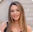 Smiling woman with straight blonde hair wearing a black top in front of a blurred background.